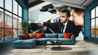 Let me show you interior of this car. Handsome young classic car salesman standing in the dealership and helping a client to make a decision about new car Wall mural